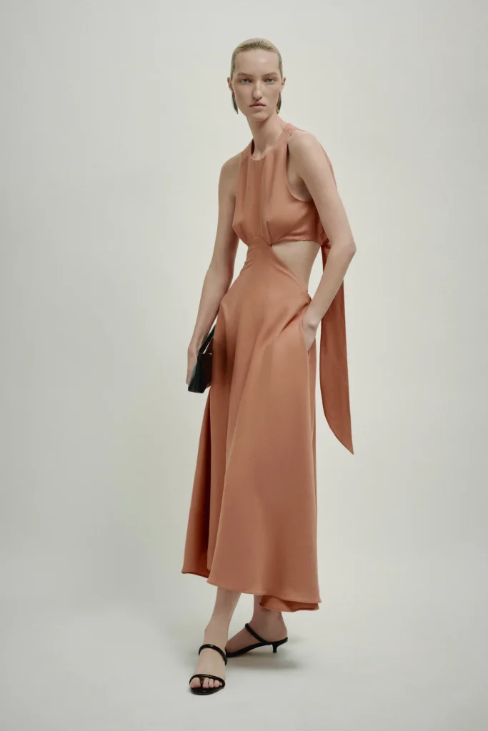 Model wearing a peach-toned sleeveless cutout gown with a draped back, holding a small black clutch and wearing black sandals in a studio.