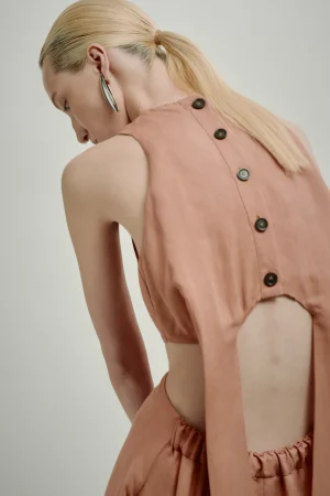Back view of a person with blonde hair in a ponytail wearing a peach sleeveless top with a buttoned back opening and exposed mid-back, wearing large silver earrings.