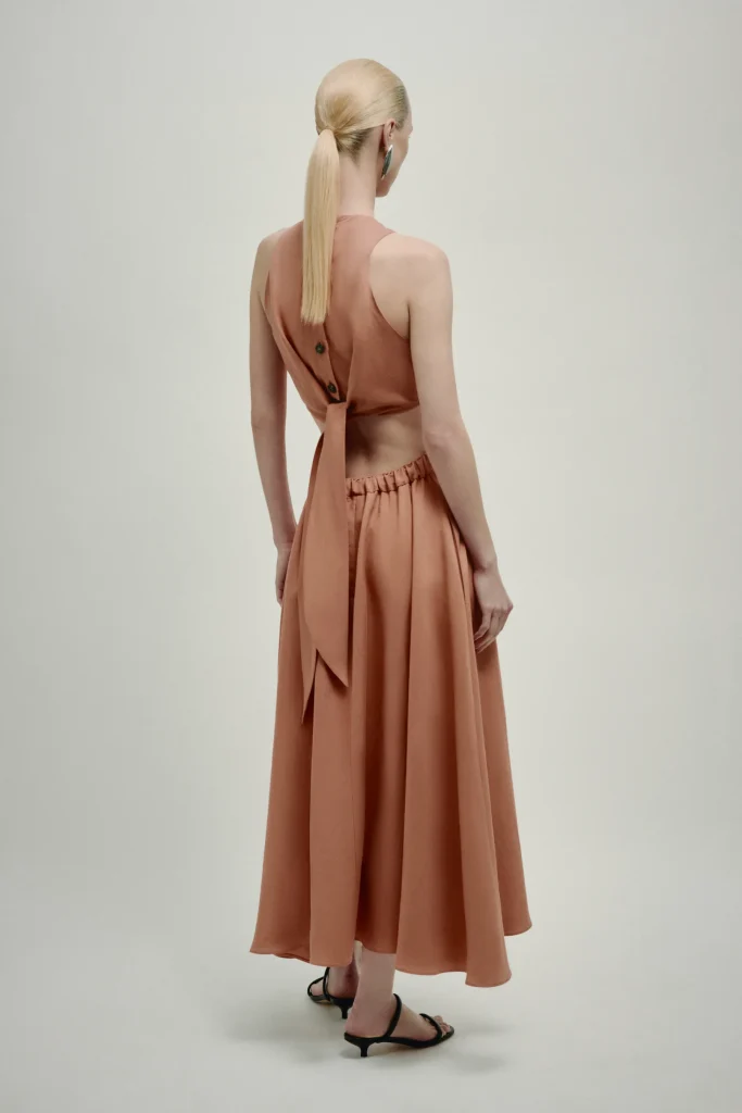 Back view of a model in a dusty rose two-piece dress with a tied back detail and long flowing skirt, wearing black sandals and a ponytail.