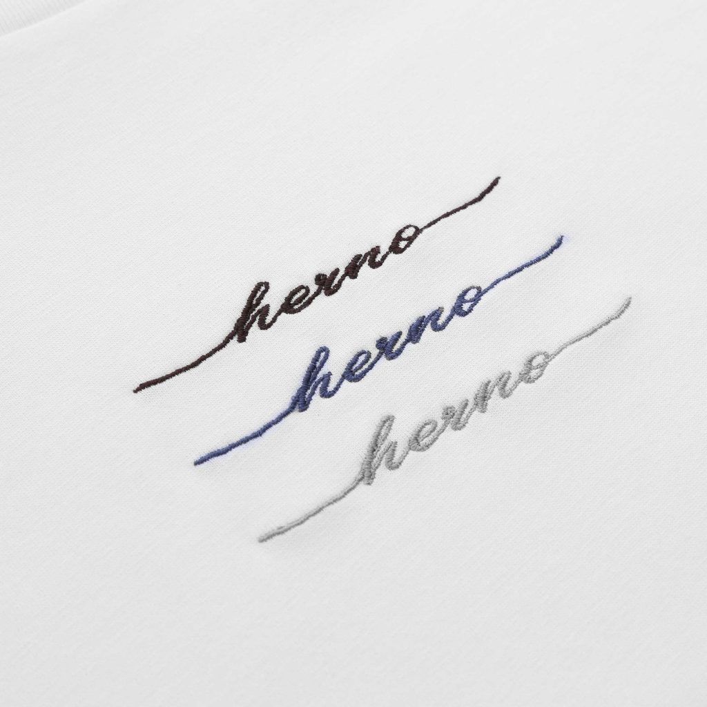 Close-up of white fabric with diagonal, multicolored cursive embroidery in brown, blue, and gray threads (partial words).