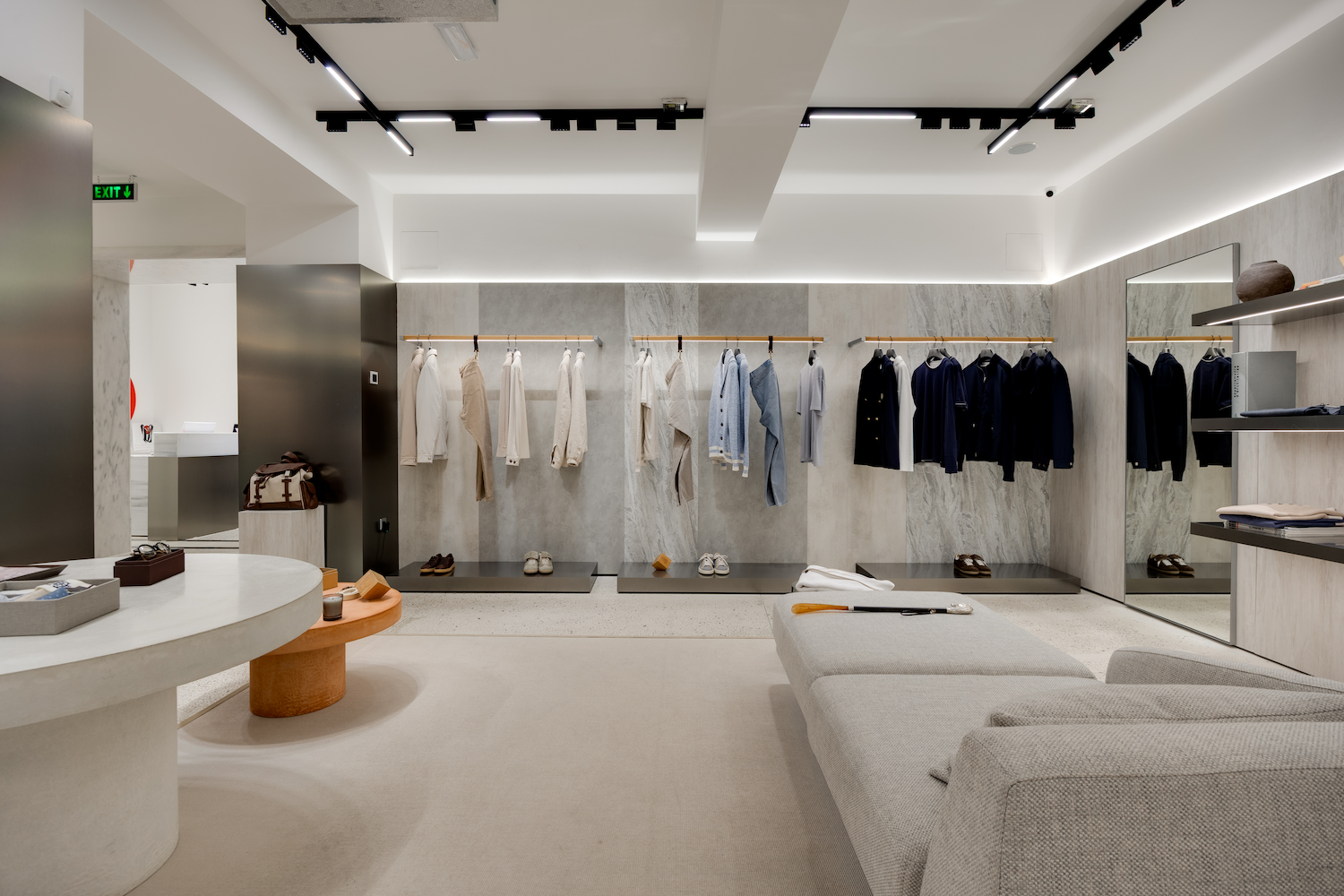 Minimalist boutique interior with hanging racks of neutral and dark clothing along a marble wall.