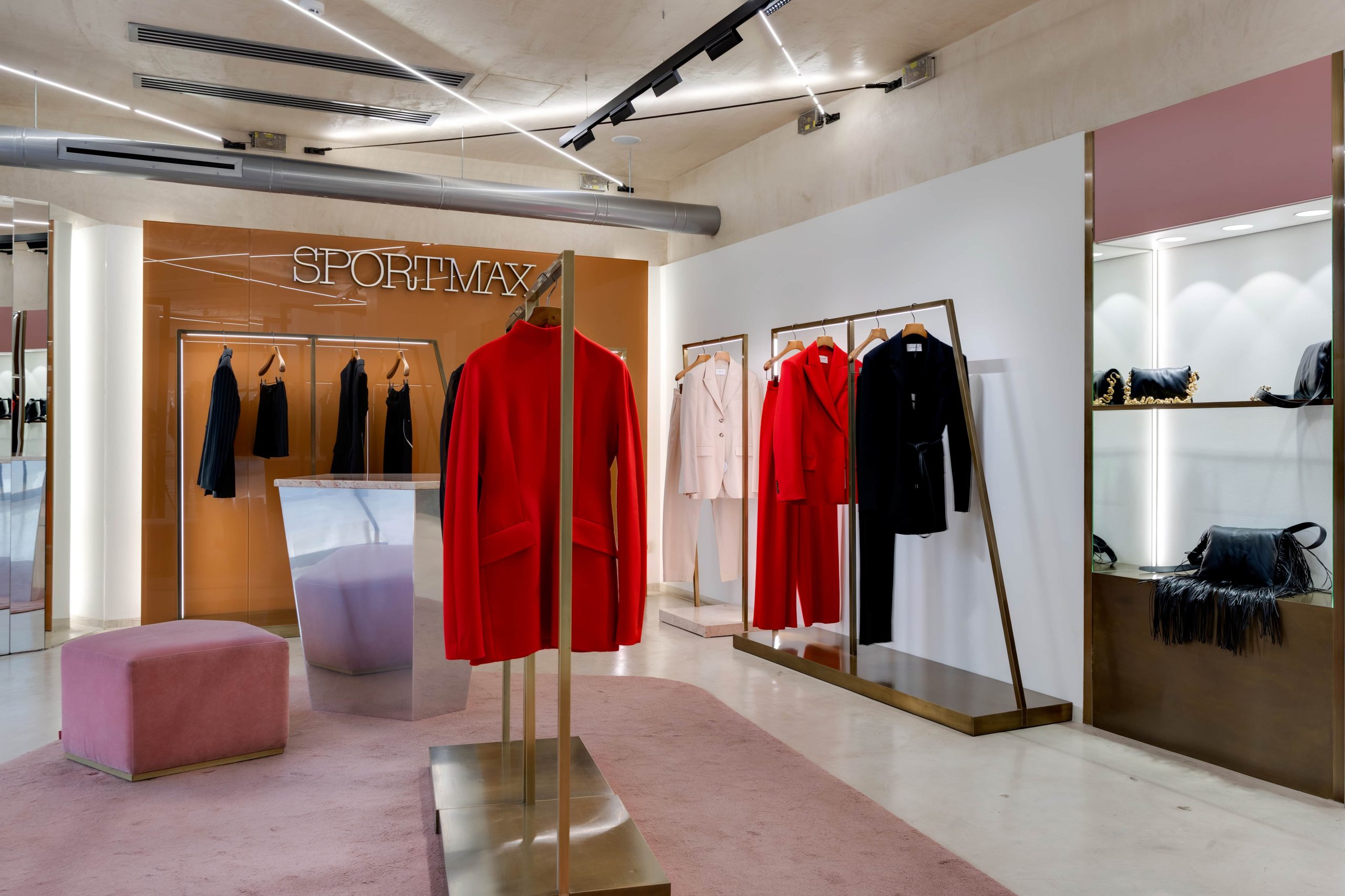 Boutique interior showing clothing on racks, bold red coats, and a glossy SPORTMAX wall in warm lighting.