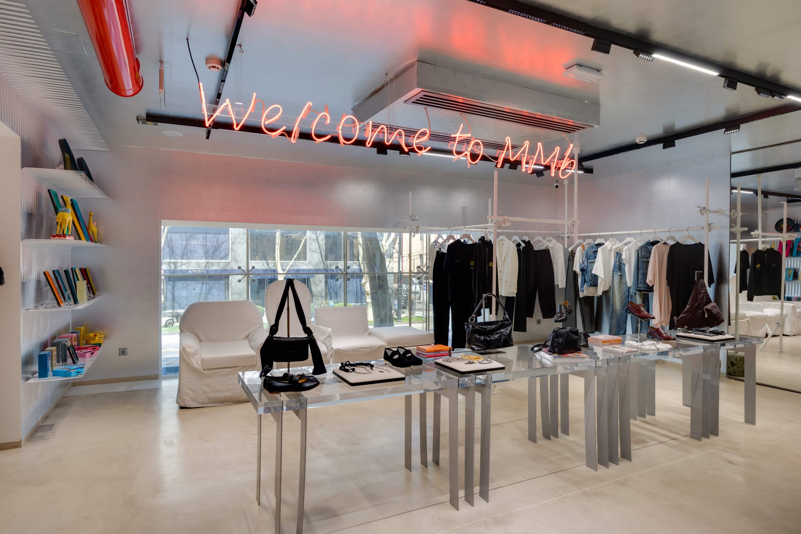 Modern clothing store interior with glass display tables, racks of shirts and dresses, and a neon 'Welcome to MMM' sign above.