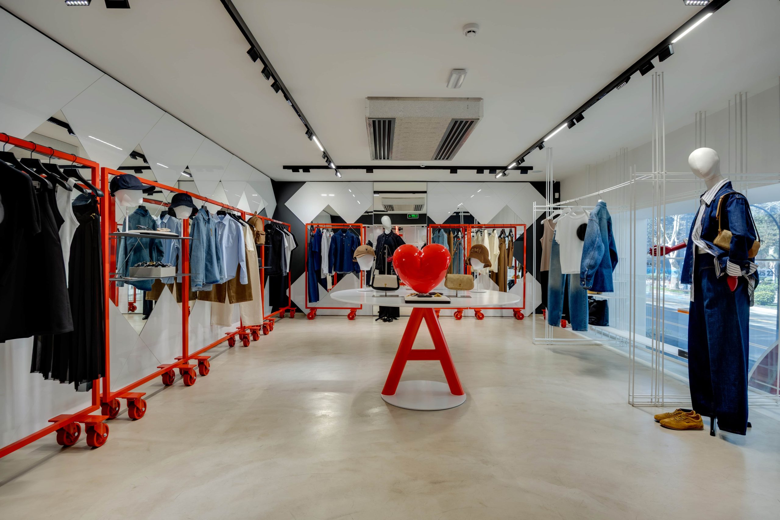 Interior of a modern clothing store with red display racks, hanging shirts and jackets, and mannequins near a white round table with red legs.