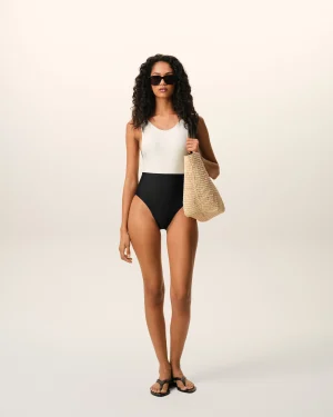 Woman in a black-and-white color-block swimsuit, sunglasses, sandals, carrying a woven straw bag against a pale background.