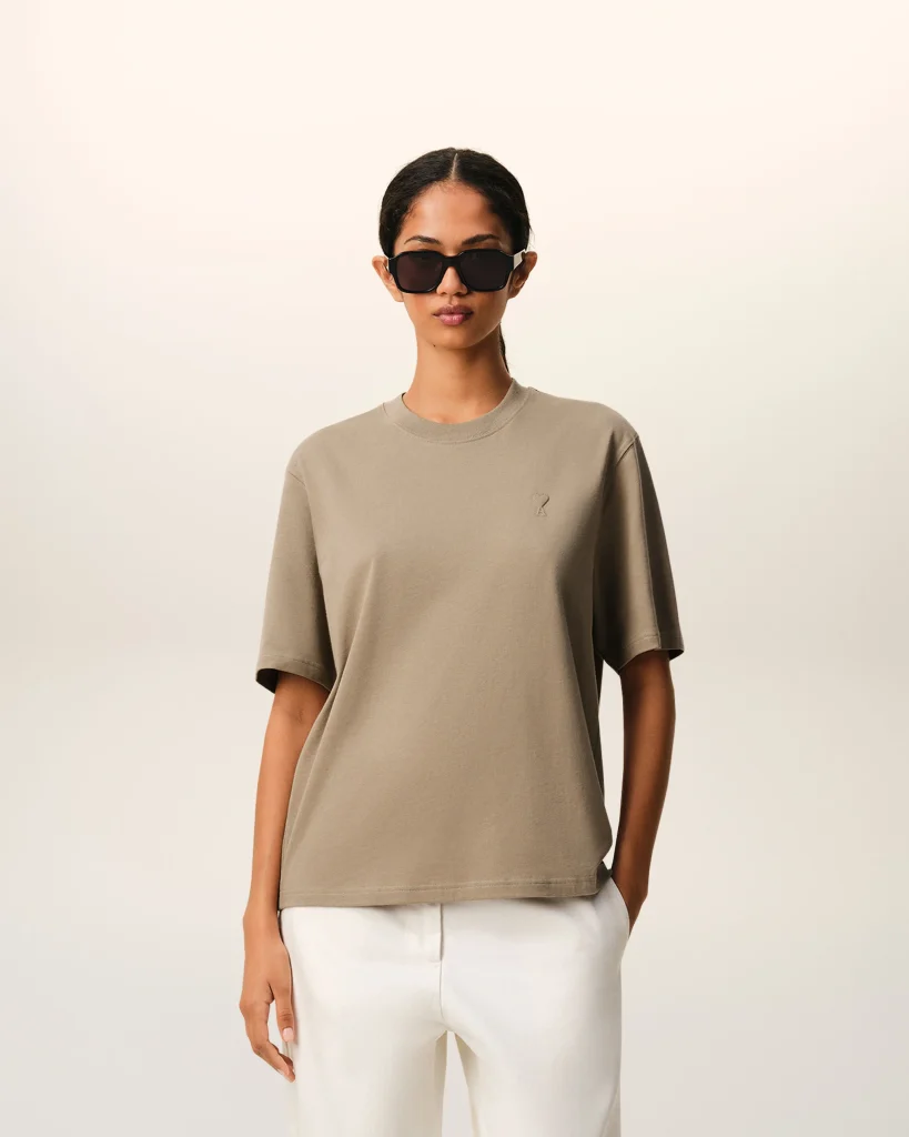 Woman wearing oversized taupe T-shirt and large black sunglasses, standing against a pale gradient background.
