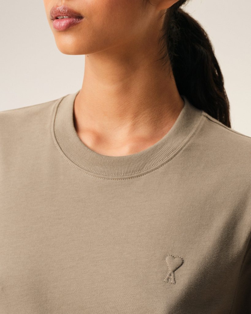 Beige crewneck sweatshirt close-up with a small stitched heart emblem on the chest.
