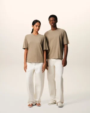 Two models stand side by side wearing oversized beige t-shirts and white pants against a pale background, showcasing a neutral look with sandals.