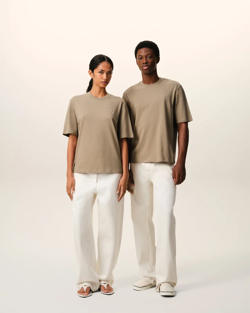 Two models stand side by side wearing oversized beige t-shirts and white pants against a pale background, showcasing a neutral look with sandals.