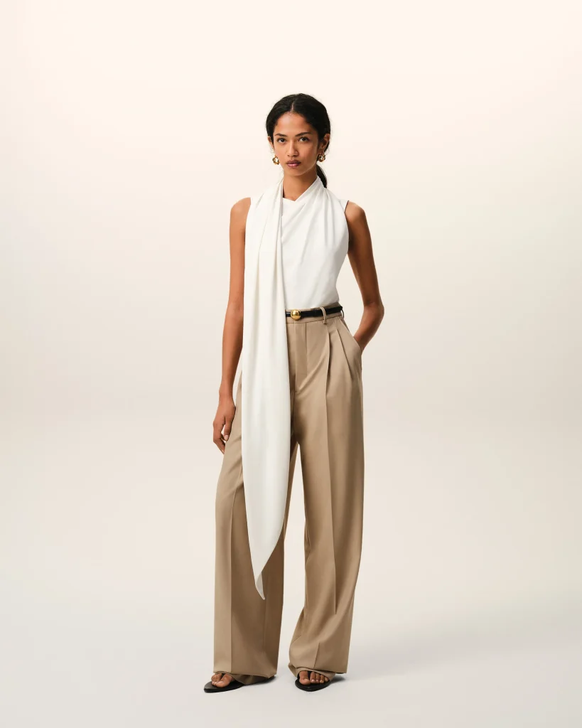 Model wearing a white draped sleeveless top with beige wide-leg trousers and black sandals, posing against a light background.