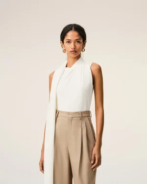 Woman wearing a white draped sleeveless top with beige high-waisted trousers and gold hoop earrings, posed against a light beige background in a studio.