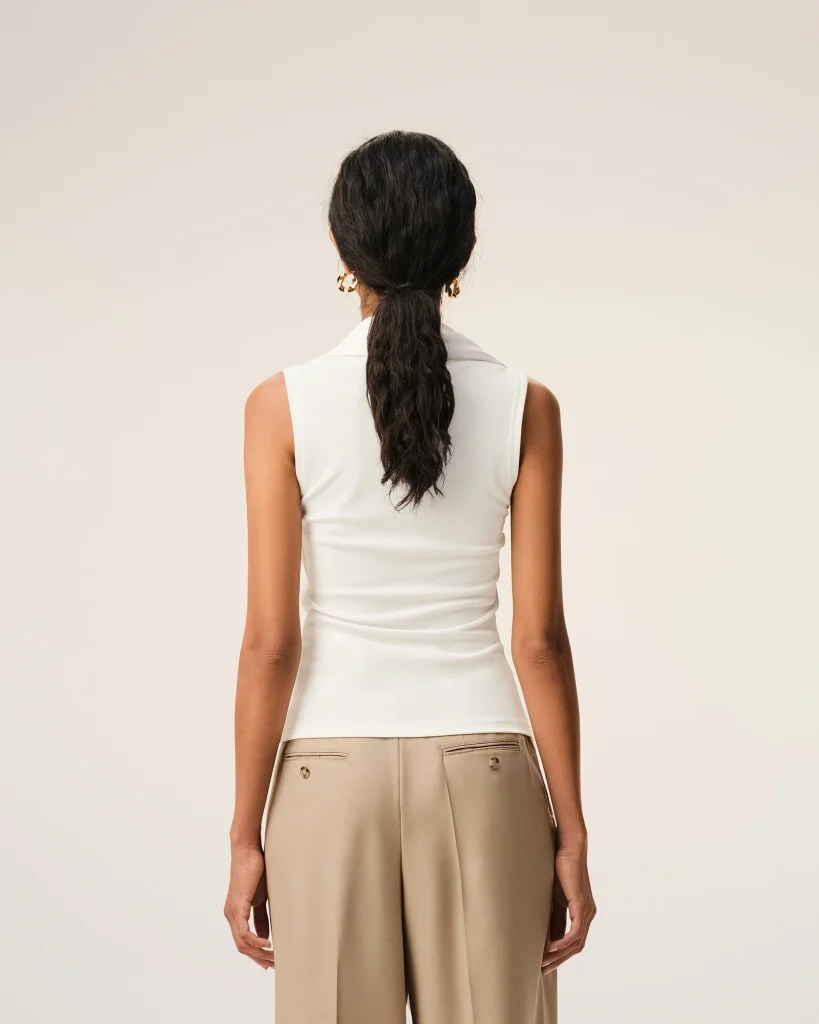 Back view of a woman in a white sleeveless top and beige wide-leg pants, with dark wavy hair in a low ponytail and gold hoop earrings.