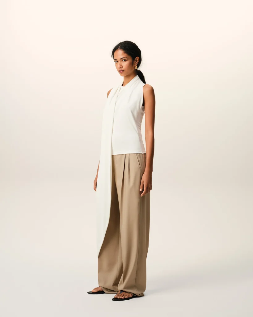 Woman in a sleeveless white top with a long scarf, beige wide-leg pants, standing barefooted with sandals, against a pale gradient backdrop.
