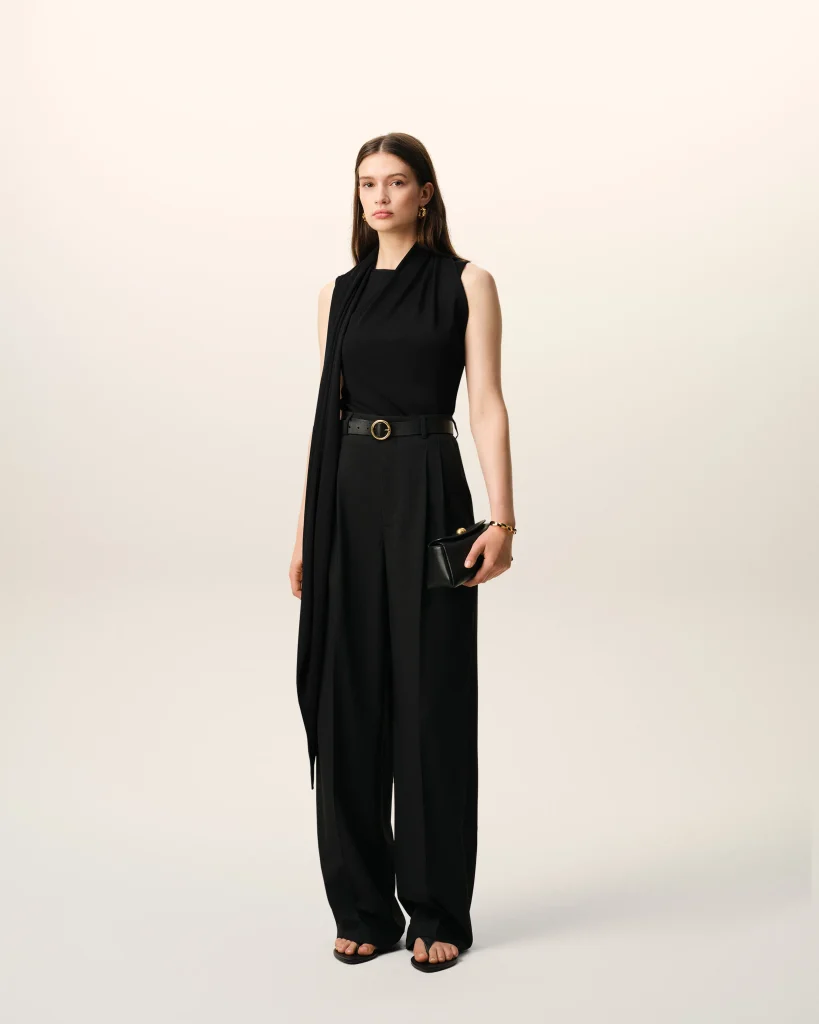Full-body portrait of a woman in a black sleeveless jumpsuit with a belt, wearing gold earrings and holding a small black clutch, standing against a light neutral backdrop.