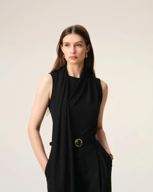 Portrait of a woman with long brown hair wearing a black draped sleeveless top and gold earrings against a beige background.