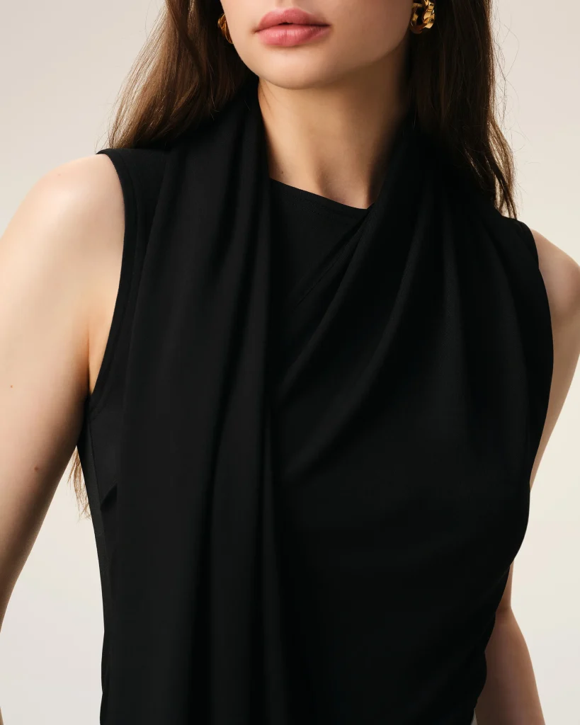Close-up of a woman wearing a black draped sleeveless top, showing neck and lips against a light beige background.
