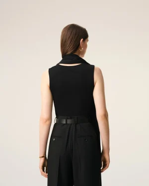 Woman viewed from behind wearing a sleeveless black top with a draped scarf and a gold hoop earring, against a light neutral background.