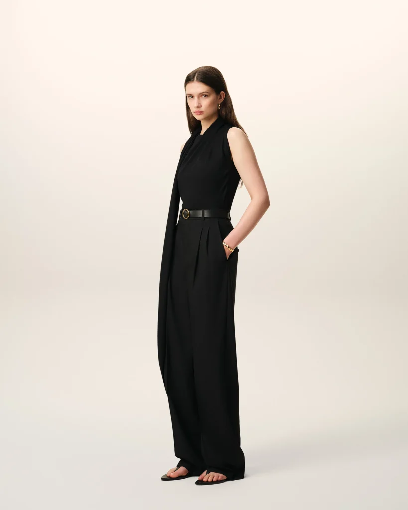 Full-body portrait of a woman in a black sleeveless outfit with a belt, standing against a pale gradient backdrop.