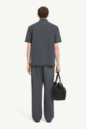 Man seen from behind wearing a loose dark gray short-sleeve shirt and matching wide-leg pants, holding a black duffel bag by the strap.