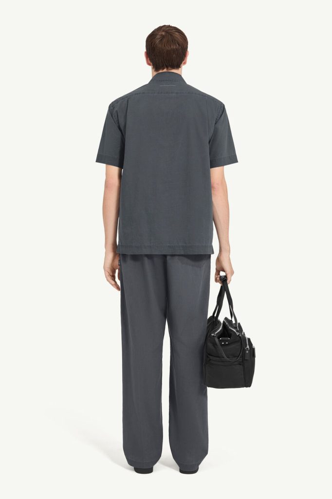 Man seen from behind wearing a loose dark gray short-sleeve shirt and matching wide-leg pants, holding a black duffel bag by the strap.
