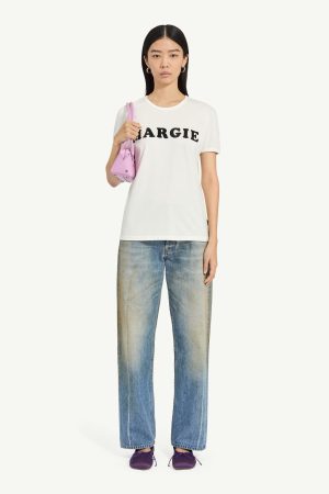 Woman in a white 'MARGIE' T-shirt, faded blue jeans, purple flats, and a pink shoulder bag coast-to-hip.