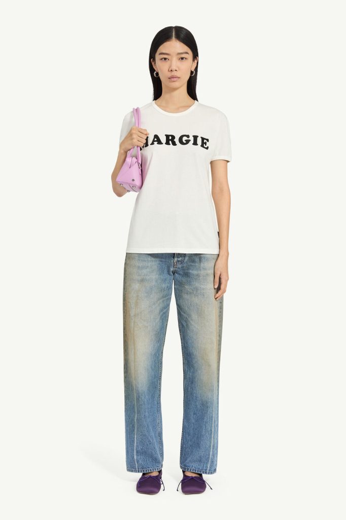 Woman in a white 'MARGIE' T-shirt, faded blue jeans, purple flats, and a pink shoulder bag coast-to-hip.