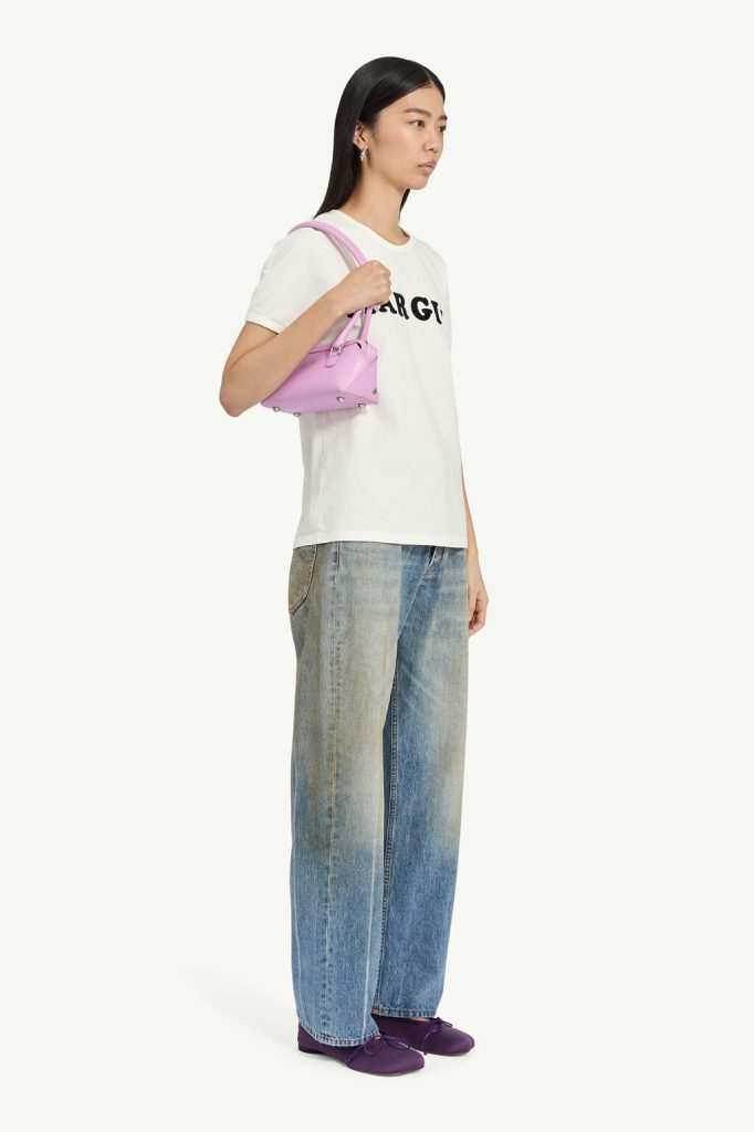 Side profile of a woman wearing a white graphic T-shirt, wide-leg jeans, purple shoes, and a pink shoulder bag.