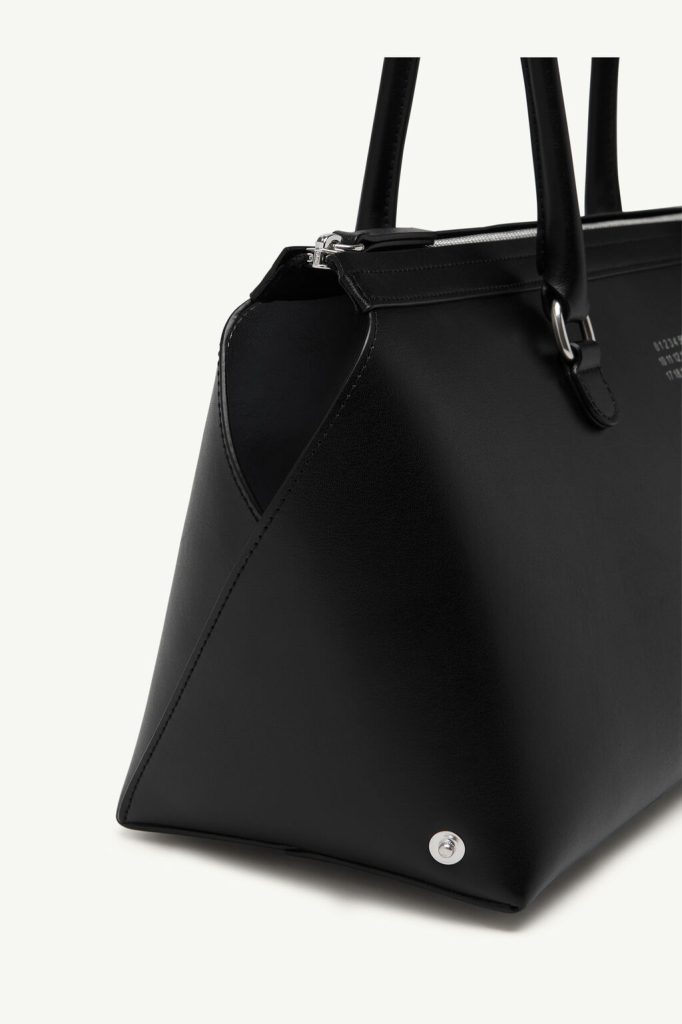 Close-up of a black leather handbag showing the corner, zipper top, handles, and a metal stud on the base.