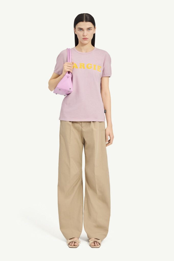 Woman wearing a pastel pink T-shirt with the word 'MARGIE' in yellow, paired with beige wide-leg pants and a pink shoulder bag.