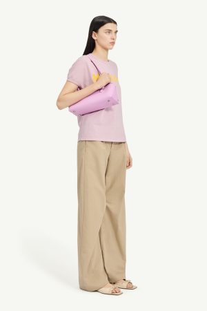 Model in pastel pink T-shirt with yellow logo, beige wide-leg pants, and pink shoulder bag; neutral background.