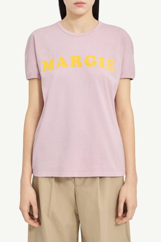 Light pink crew-neck T-shirt with the yellow word 'MARGIE' across the chest, worn by a model in beige pants (informative product image).