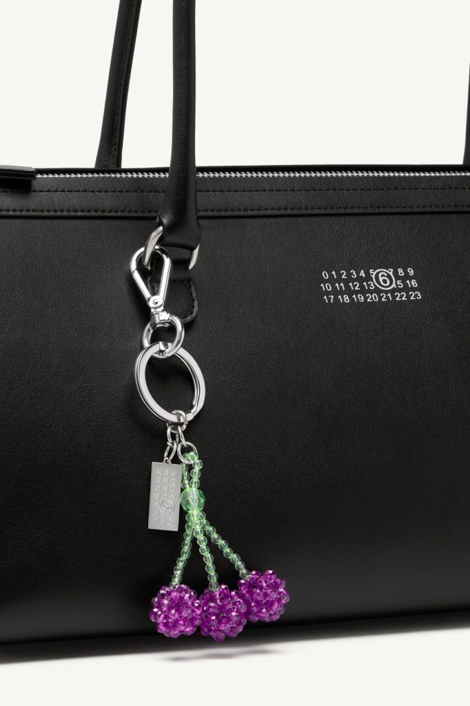 Close-up of a black leather handbag with a silver keychain charm featuring green bead strands and purple bead clusters hanging from the ring near the zipper.