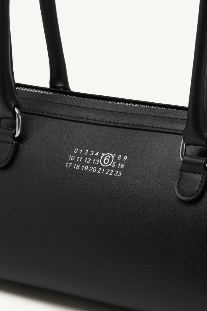 Close-up of a black leather handbag with a white calendar-style emblem and circular logo near the zipper.