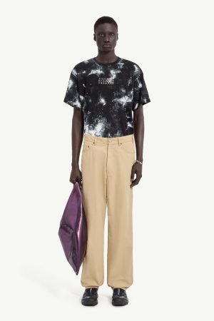 Male model in a black and white tie-dye T-shirt with beige wide-leg pants, holding a purple clutch.