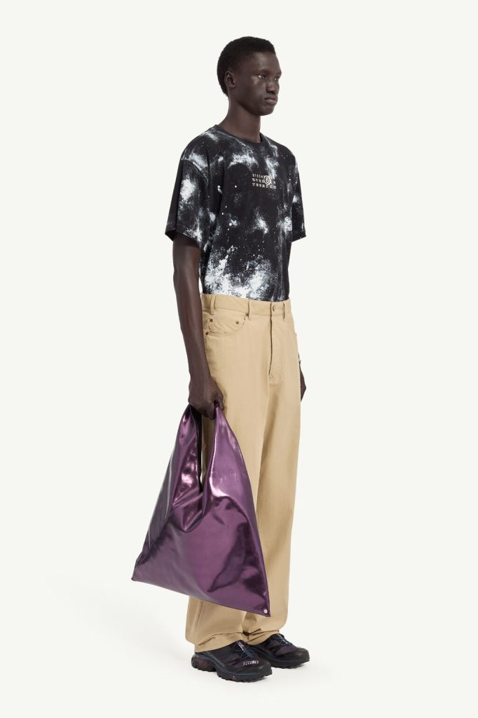 Full-body shot of a Black man wearing a black splatter-print T-shirt, beige wide-leg pants, dark sneakers, and holding a large shiny purple tote bag.
