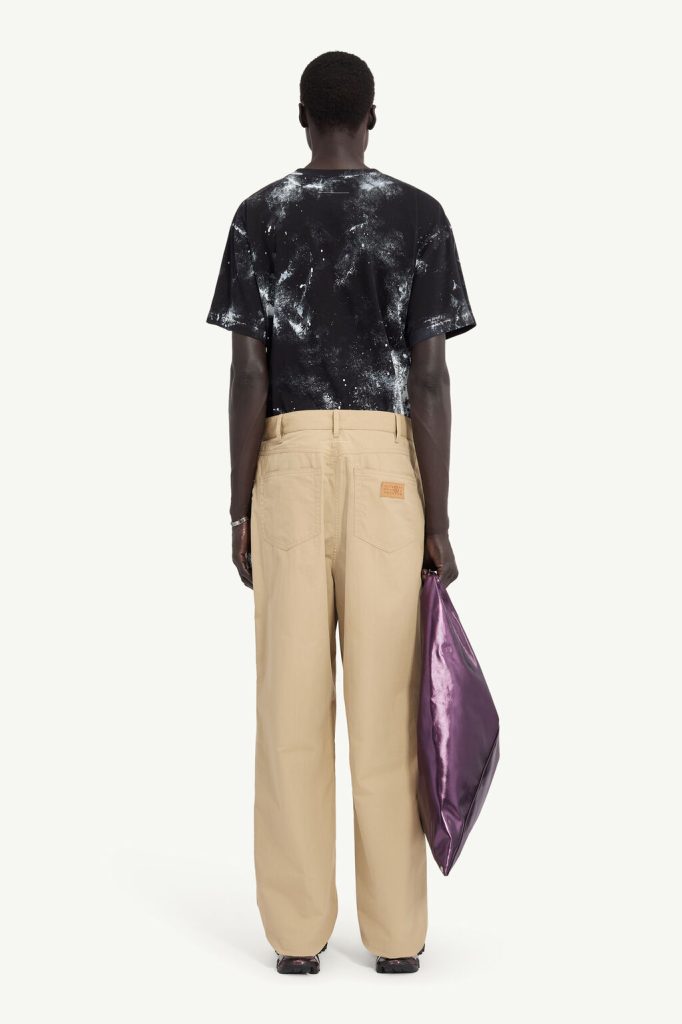 Back view of a person in a black splatter-print T-shirt and beige wide-leg pants, holding a metallic purple bag.