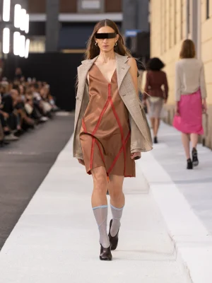 Model walks a runway in a bronze satin dress with red seam lines and a light plaid blazer draped over shoulders; sheer knee-high socks and dark heels.