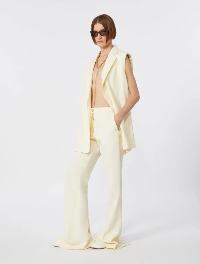 Model wearing a light cream sleeveless suit with wide-leg pants, sunglasses, and gold jewelry.
