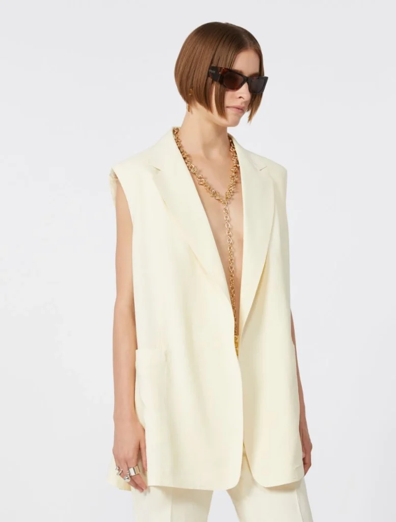 Model wearing a cream sleeveless blazer with matching pants, large dark sunglasses, and a long gold chain necklace.