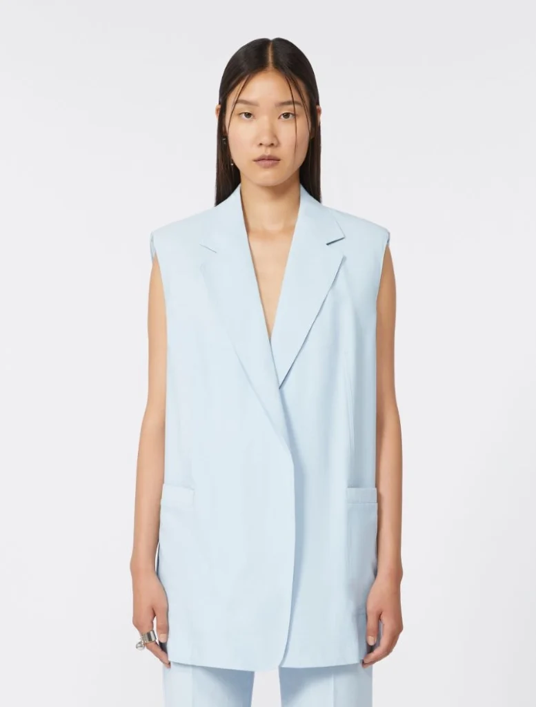 Woman wearing an oversized light blue sleeveless blazer with long lapels and front pockets in a studio.