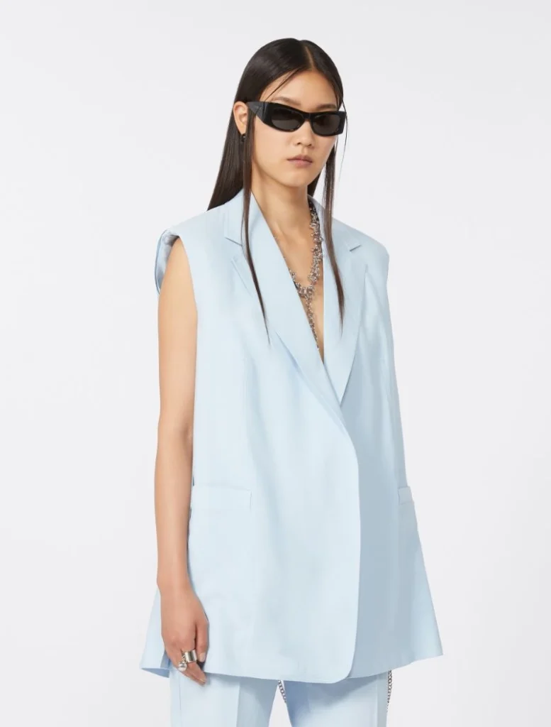 Model wearing a pale blue sleeveless blazer with matching trousers, black rectangular sunglasses, and a chunky silver necklace.