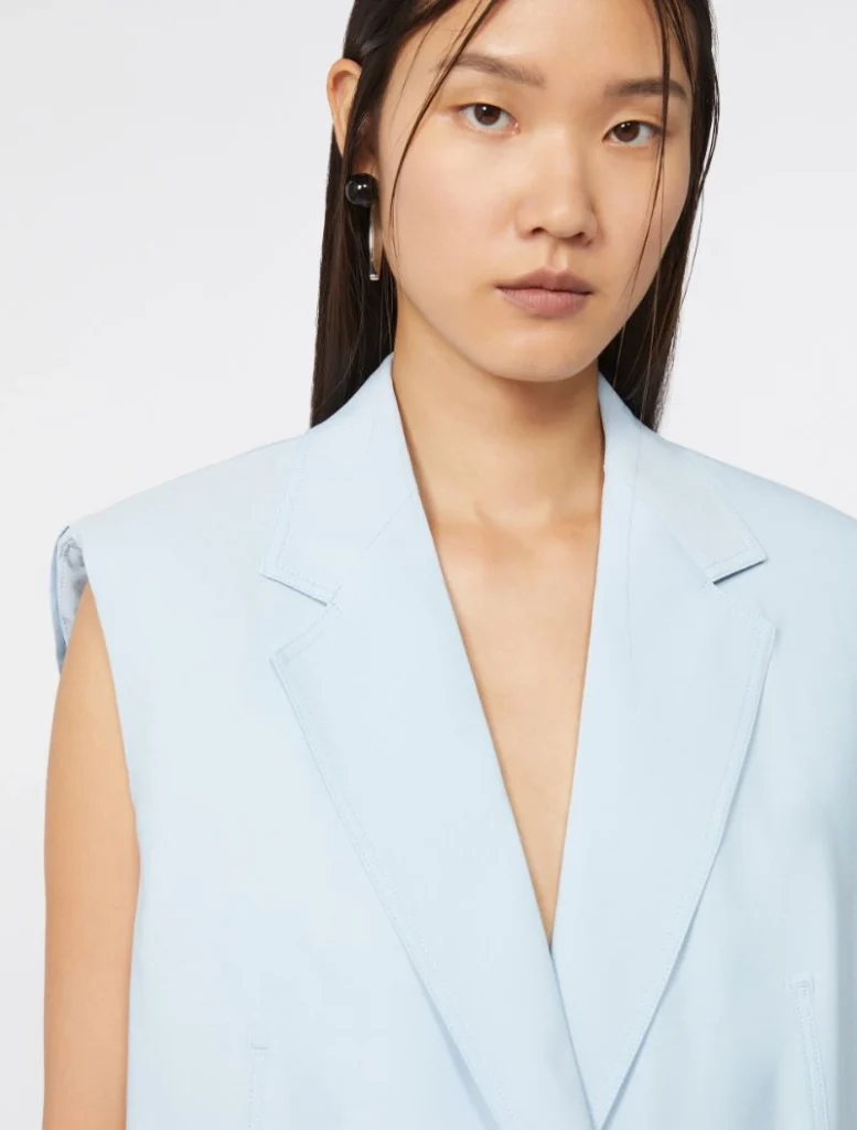 Model wearing a light blue sleeveless blazer, facing the camera in a studio setting.