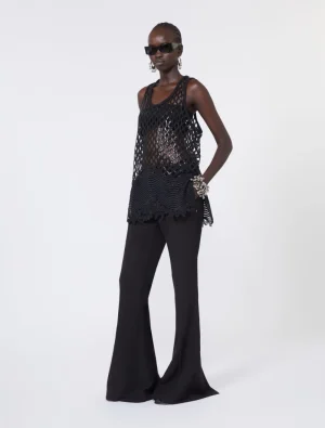 Model wearing a black mesh tank top over a black bralette, with wide-leg black trousers, sunglasses, and chunky jewelry in a studio pose