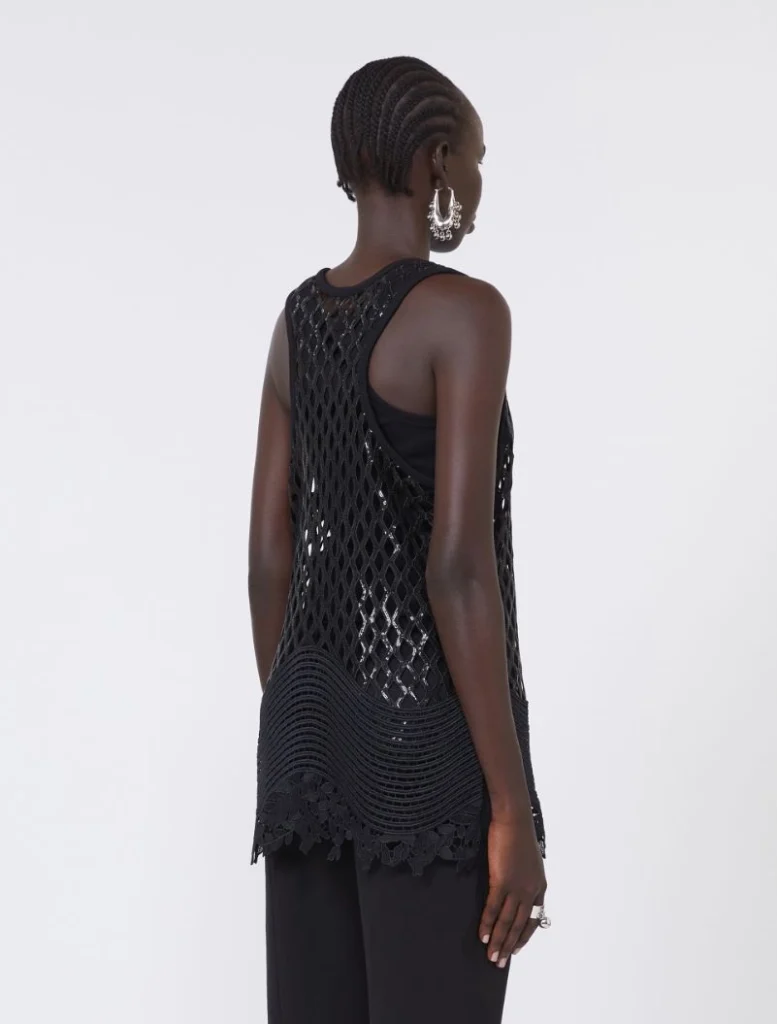 Back view of a model wearing a black sleeveless crochet top with a lattice pattern over a black tank, paired with bold silver earrings and black pants, standing against a light background.