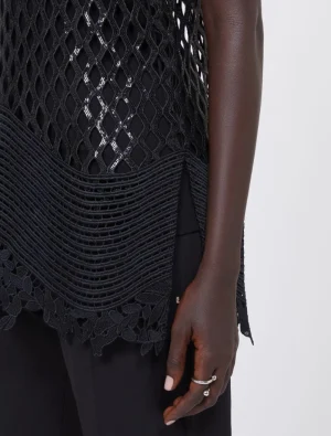 Close-up of a person wearing a black mesh top with a wavy, layered hem and floral lace trim, showing their hand with rings.