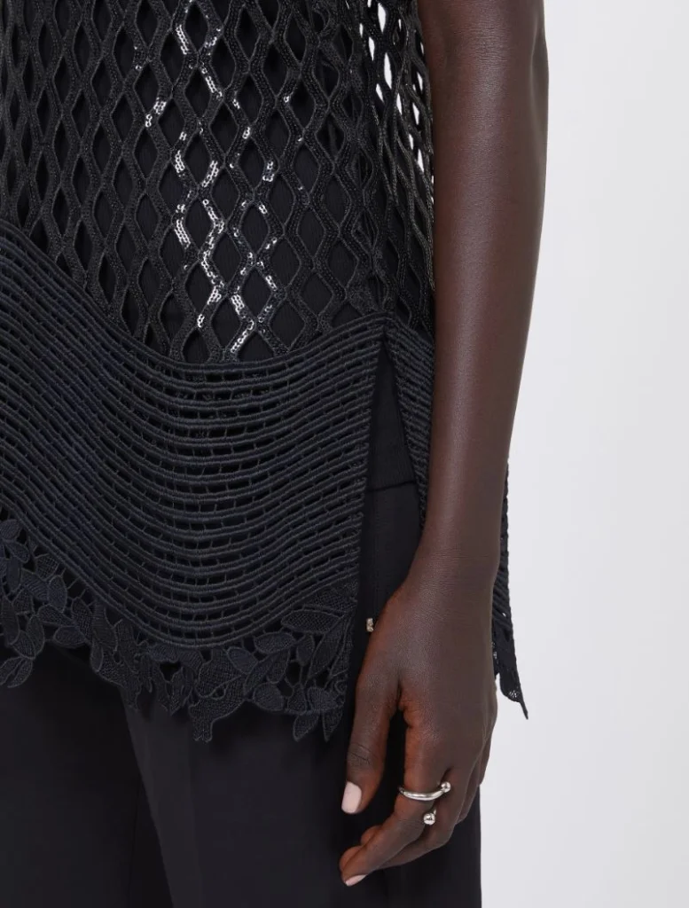 Close-up of a person wearing a black mesh top with a wavy, layered hem and floral lace trim, showing their hand with rings.