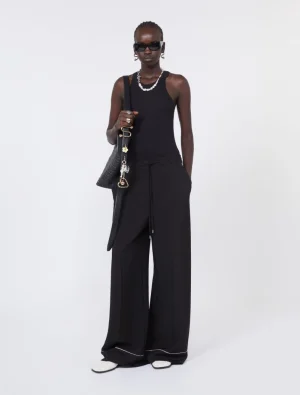 Model in a black sleeveless top and wide-leg pants, carrying a black shoulder bag with pearls, sunglasses, and white sneakers.