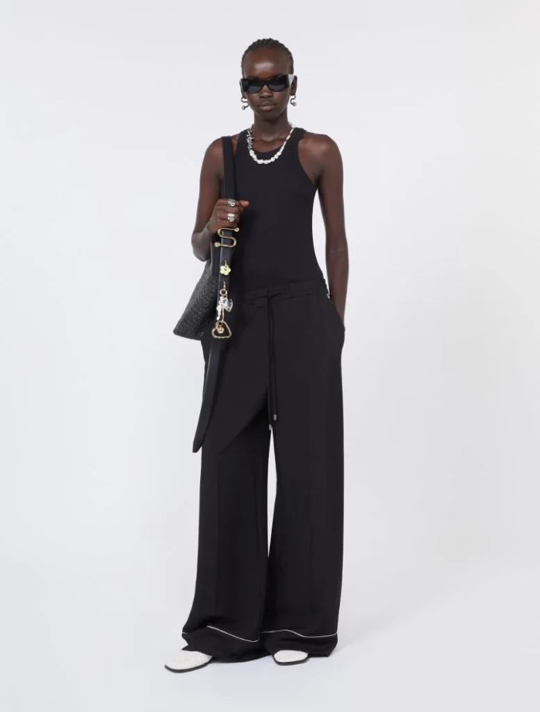 Model in a black sleeveless top and wide-leg pants, carrying a black shoulder bag with pearls, sunglasses, and white sneakers.