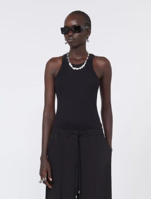 Fashion model wearing a black tank top, black pants, pearl necklace, and dark sunglasses, standing against a neutral backdrop.