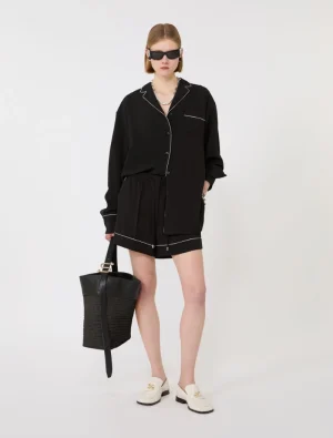 Model wearing a black pajama-style shirt and shorts with white piping, cream loafers, oversized sunglasses, and a black woven tote bag.